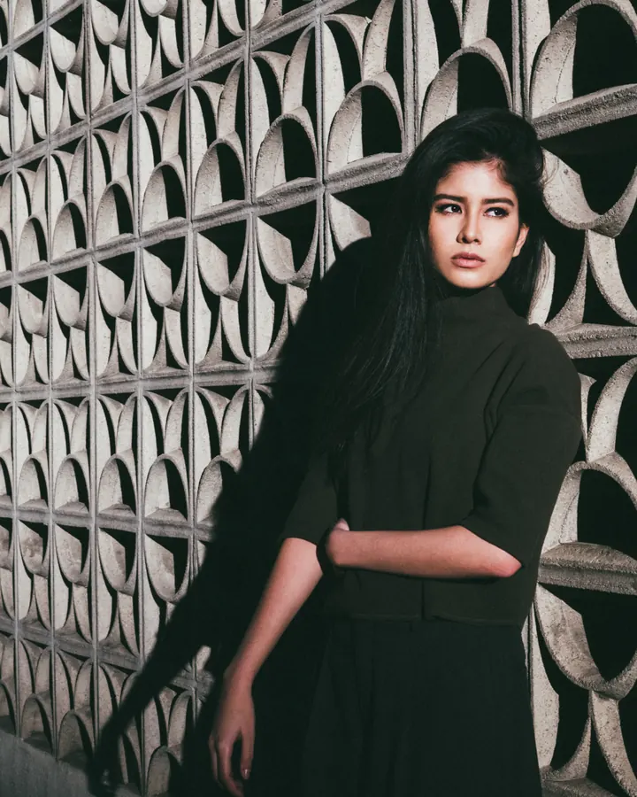 Femme en tenue noire, debout devant un mur décoratif, bras croisés, regard intense.