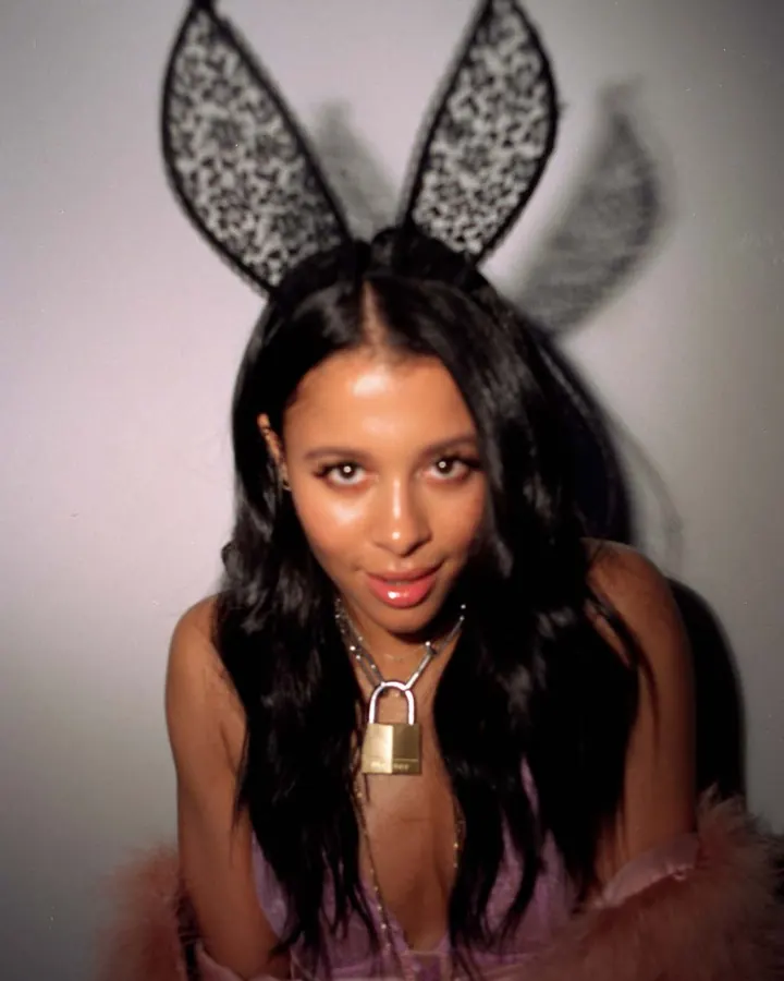 Femme aux cheveux noirs, portant des oreilles de lapin et un collier avec un cadenas, vêtue d'une robe rose et d'une écharpe fourrée.
