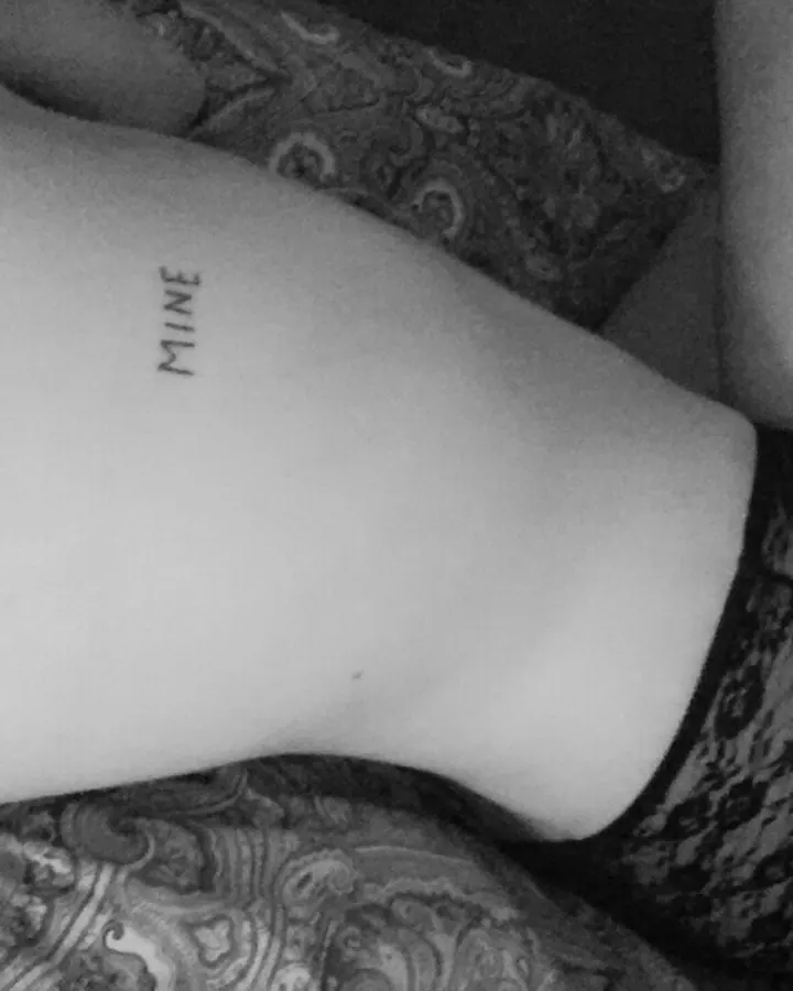 Femme debout, portant un haut noir et un jean bleu, les mains sur ses seins, tenant un téléphone doré pour se prendre en photo, avec un collier et une montre blanche.